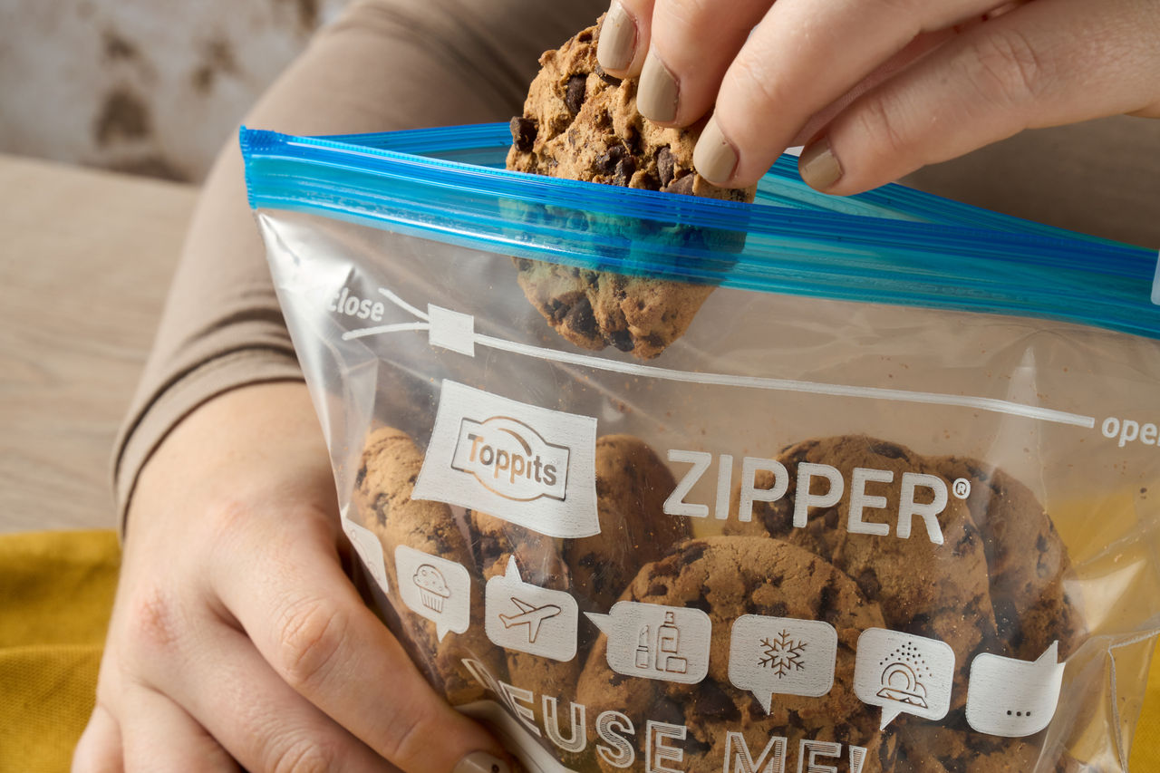 Eine Hand nimmt einen Schoko-Cookie aus einem Toppits Zipper Beutel auf einem Holztisch mit gelbem Tuch.