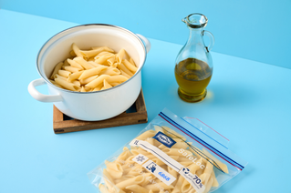 Ein weißer Topf mit gekochter Pasta steht auf einem Holzuntersetzer, daneben eine Glasflasche mit Öl und ein Gefrierbeutel mit Penne auf einem hellblauen Untergrund. 