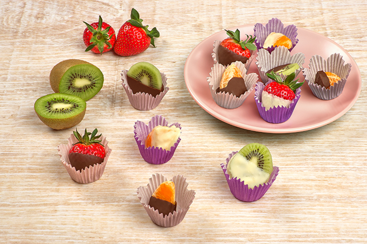 Fruchtstücke wie Erdbeeren, Kiwis und Mandarinen, teilweise mit Schokolade überzogen, in bunten Toppits Papierförmchen und auf einem rosa Teller angerichtet. 