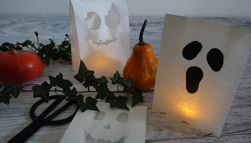 Zwei leuchtende Halloween-Lichttüten aus Papier mit ausgeschnittenen Geister- und Kürbisgesichtern stehen auf einem Holztisch. Daneben liegen ein Zierkürbis, ein roter Kürbis, Efeuranken und eine Schere